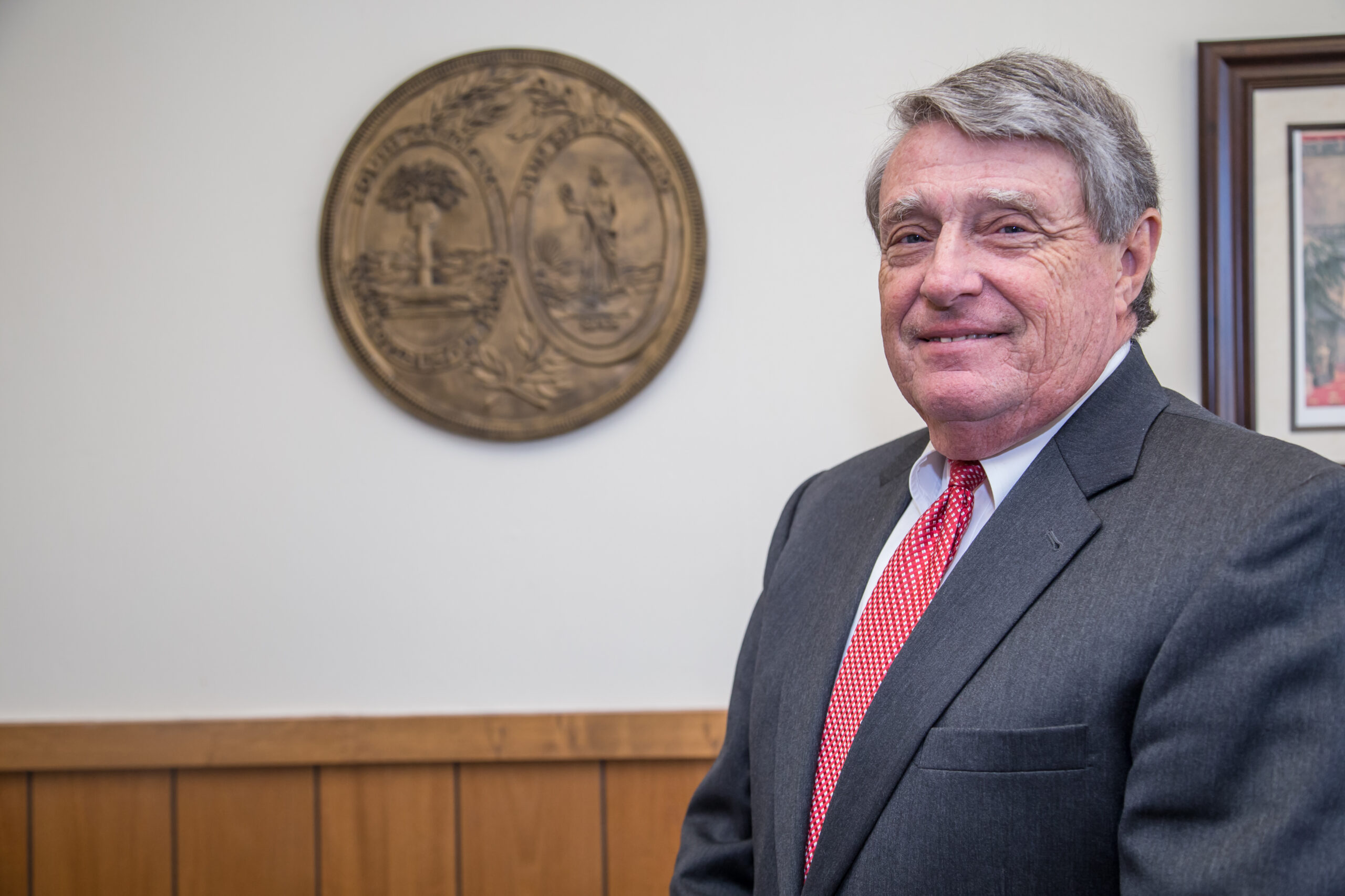 Magill stands, smiling, in front of the South Carolina seal in a gray suit and red tie.