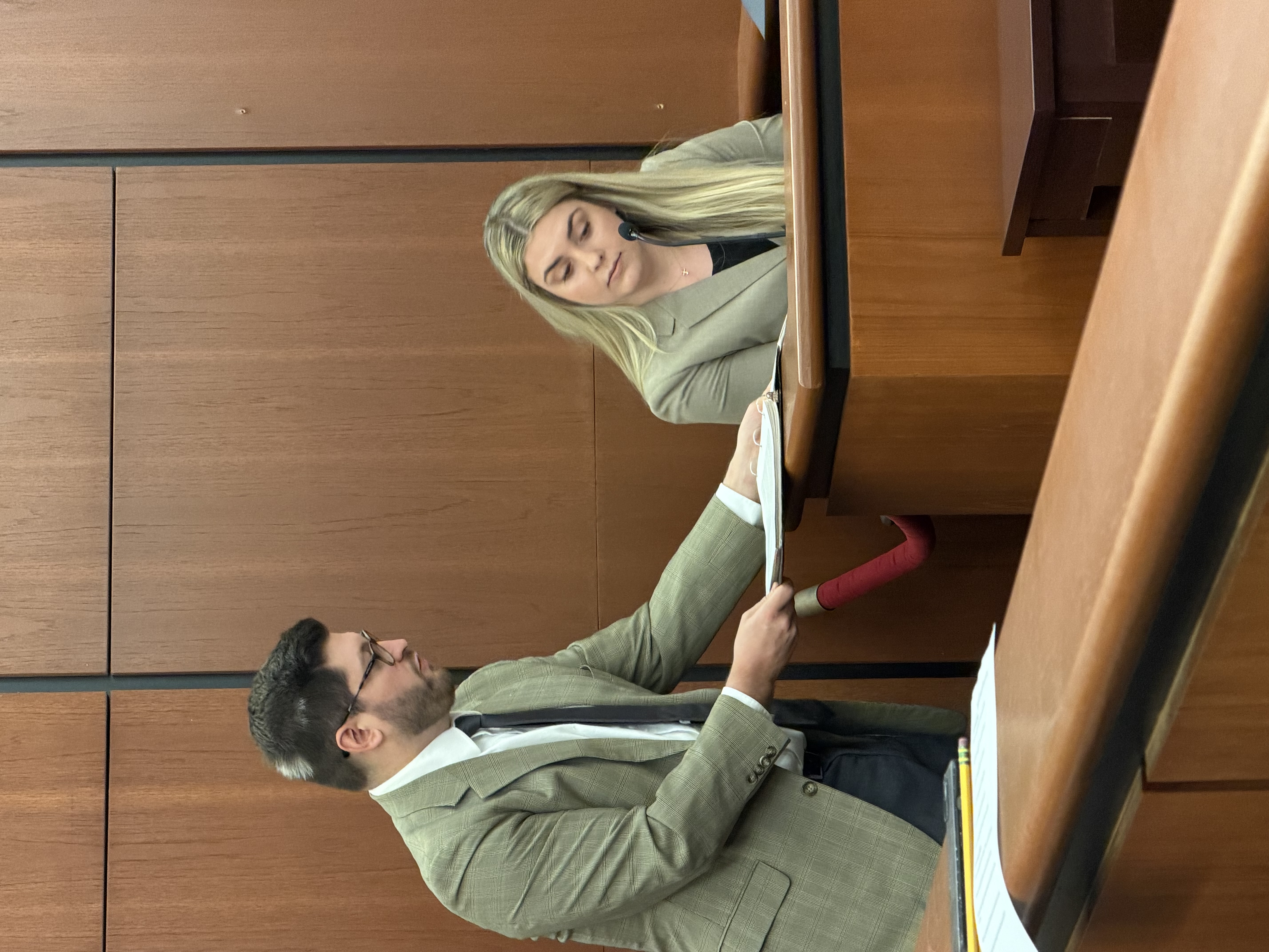 Photo of a woman sitting in a courtroom witness box. A man standing to her right is showing her a document.